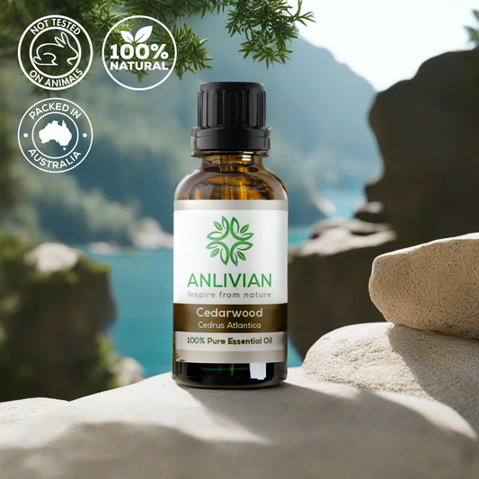 bottle of Anlivian Cedarwood (Cedrus Atlantica) 100% Pure Essential Oil with a black cap. The bottle is placed on a light-colored rock with a blurry, beautiful natural background of water, mountains, and greenery. Overlaid icons indicate the product is Not Tested on Animals, 100% Natural, and Packed in Australia.